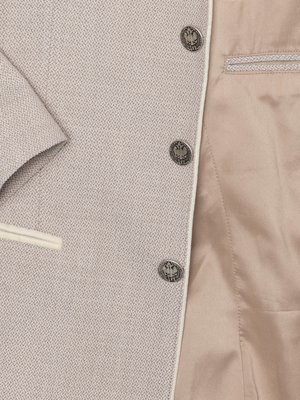 Nahaufnahme einer hellgrauen Jacke mit weißen Ziernähten und drei Knöpfen. Innenfutter in beige. Die Knöpfe zeigen ein geprägtes Emblem.  Die Jacke hat eine kleine Brusttasche. Der Ärmelansatz ist links sichtbar.