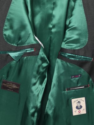 Nahaufnahme des Innenfutters einer grauen Herrenjacke mit grünen Seidenfutter. Auf dem Futter befinden sich zwei Etiketten, auf denen "Vintage Barberis" und ein weiteres mit einem Cartoon-Mann abgebildet sind. Die Innentaschen haben rote und schwarze Akzente.