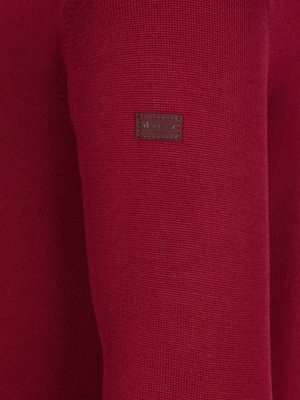 Nahaufnahme eines roten Pullovers. Auf dem linken Ärmel befindet sich ein kleines, rechteckiges braunes Etikett mit weißer Schrift. Die Struktur des Pullovers ist erkennbar.