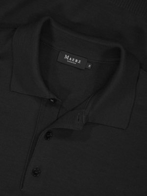 Nahaufnahme eines schwarzen Poloshirts mit Kragen und Knöpfen. Das Etikett mit dem Namen "Maerz Muenchen" ist sichtbar. Das Hemd liegt gefaltet. Die Knöpfe sind geschlossen. Die Oberfläche ist leicht strukturiert.
