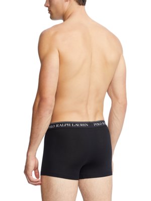Ein Mann, von hinten gesehen, trägt schwarze Boxershorts. Der Bund hat eine silberfarbene Linie und den weißen Schriftzug "POLO RALPH LAUREN". Er hat helle Haut und steht vor hellem Hintergrund.