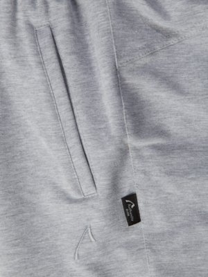 Nahaufnahme eines grauen Kleidungsstücks, wahrscheinlich einer Hose, mit einer vertikalen Tasche auf der linken Seite. Auf der rechten Seite ist ein schwarzes Etikett mit weißer Schrift zu sehen. Unterhalb des Etiketts ein kleiner, gestickter Pfeil.