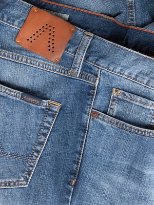 Nahaufnahme der Rückseite einer Jeans in Blau.  Zu sehen sind die Gesäßtaschen, der Bund mit einem Leder-Label. Das Label hat eine Perforierung in Form des Buchstaben "A". Die Nahtfarbe ist orange.