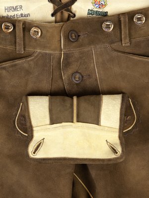 Nahaufnahme einer braunen Lederhose mit Knopfleiste, aufgesetzter Tasche und Hosenträgern. Das Leder ist leicht strukturiert. Auf dem Beleg ist eine Stickerei zu sehen. In der oberen Ecke befindet sich ein Aufnäher mit Schriftzug.