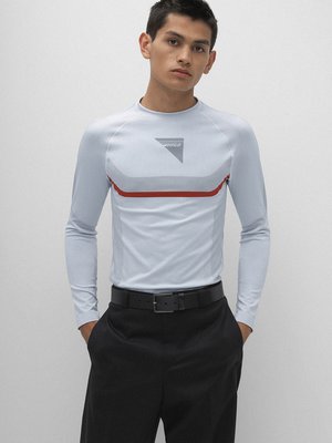 Ein junger Mann steht vor hellgrauem Hintergrund und blickt in die Kamera. Er trägt ein hellblaues Langarmshirt mit rotem Streifen und schwarzer Hose. Auf dem Shirt ist das Logo von Hugo. Seine Hände sind in den Hosentaschen.