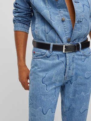 Ein Mann trägt eine blaue Jeansjacke und eine Jeanshose mit einem Muster. Er hat einen schwarzen Ledergürtel mit einer silbernen Schnalle um die Taille. Das Outfit ist aus Denim. Seine Arme und Beine sind teilweise sichtbar.