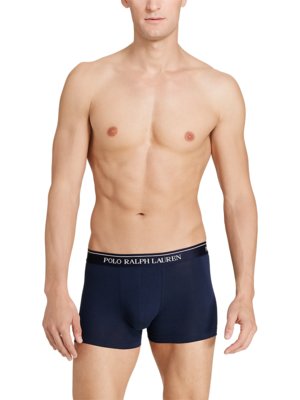 Ein Mann steht vor weißem Hintergrund, Oberkörper frei, trägt dunkelblaue Boxershorts. Auf dem Bund steht in weißer Schrift: POLO RALPH LAUREN. Der Mann schaut geradeaus.