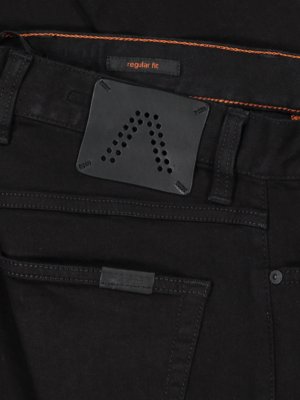 Nahaufnahme einer schwarzen Jeanshose. Auf dem hinteren Bund befindet sich ein Lederpatch mit einer Reihe von Löchern in Pfeilform. Über dem Patch steht "regular fit". Nahtdetails in Orange sind sichtbar.